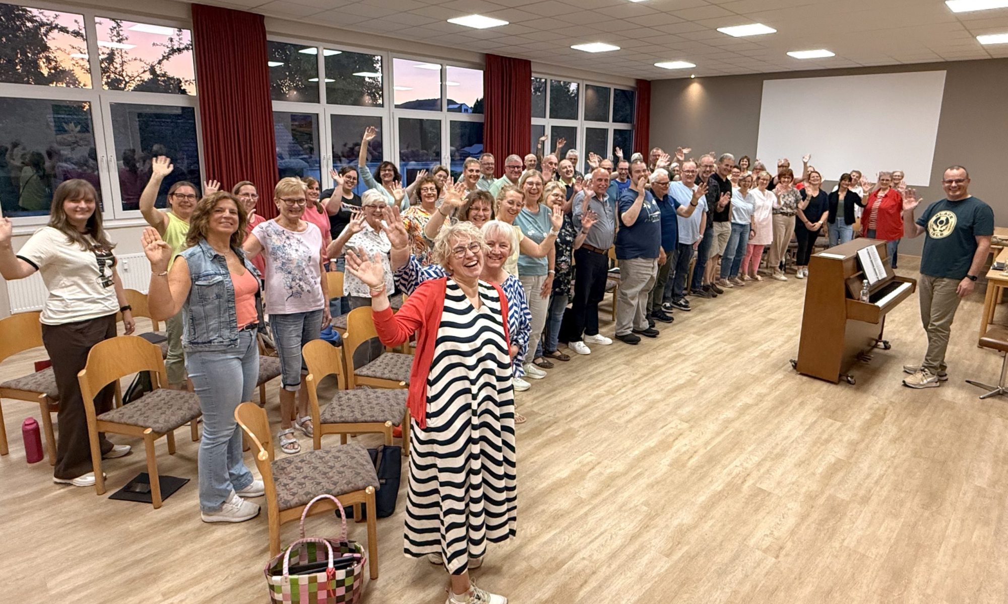 Das Bild zeigt den Chor in ihrem Probenraum Es ist von der Seite fotografiert. Alle gucken und winken freundlich in die Kamera.